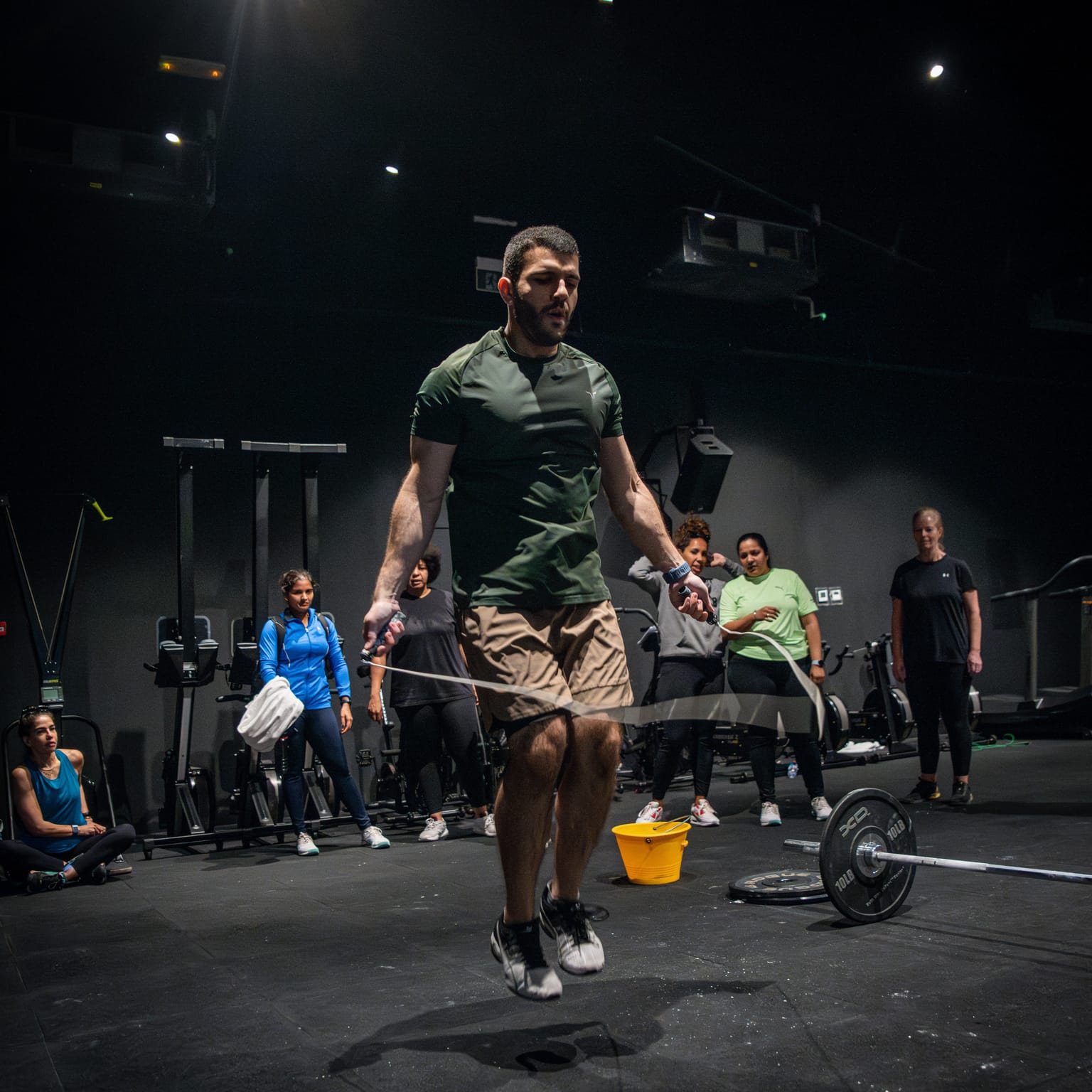 A man in a green athletic shirt performing double-unders with a jump rope in a dimly lit gym.