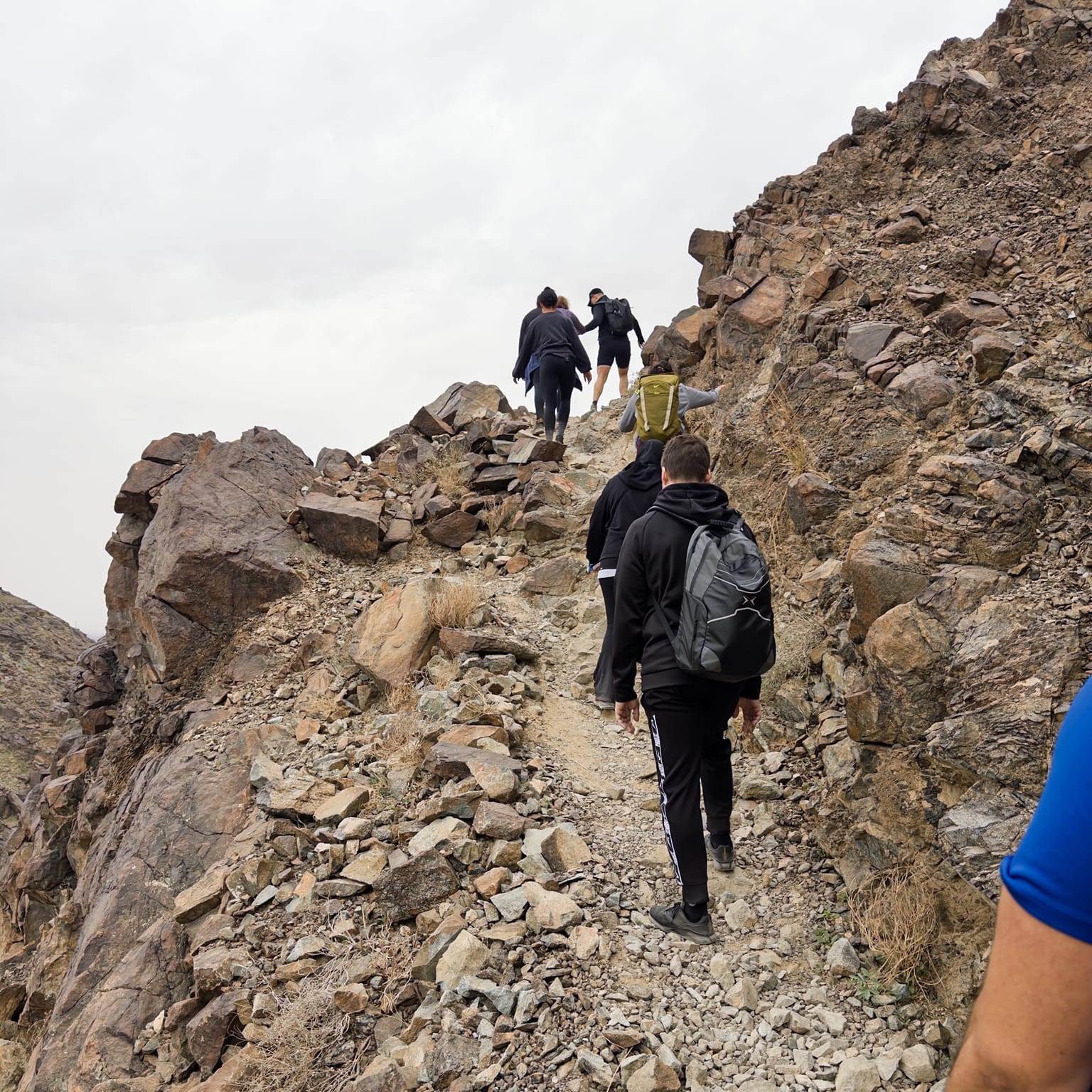 A line of hikers with backpacks ascending a steep, rocky mountain trail under a grey sky.