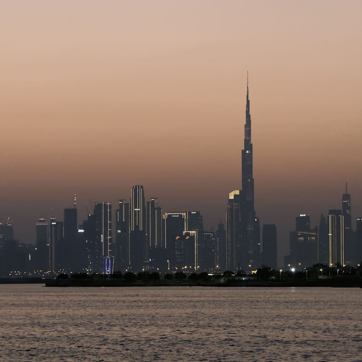 The Dubai city skyline at sunset, featuring the Burj Khalifa silhouetted against a hazy orange horizon over calm water.